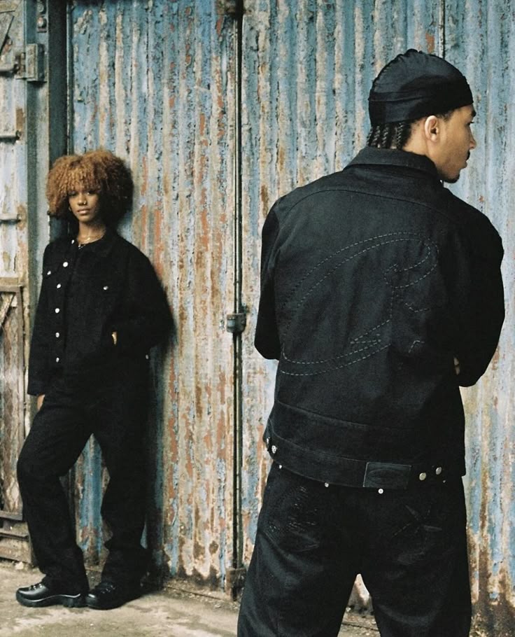 Two models wearing black denim against a weathered blue wall.